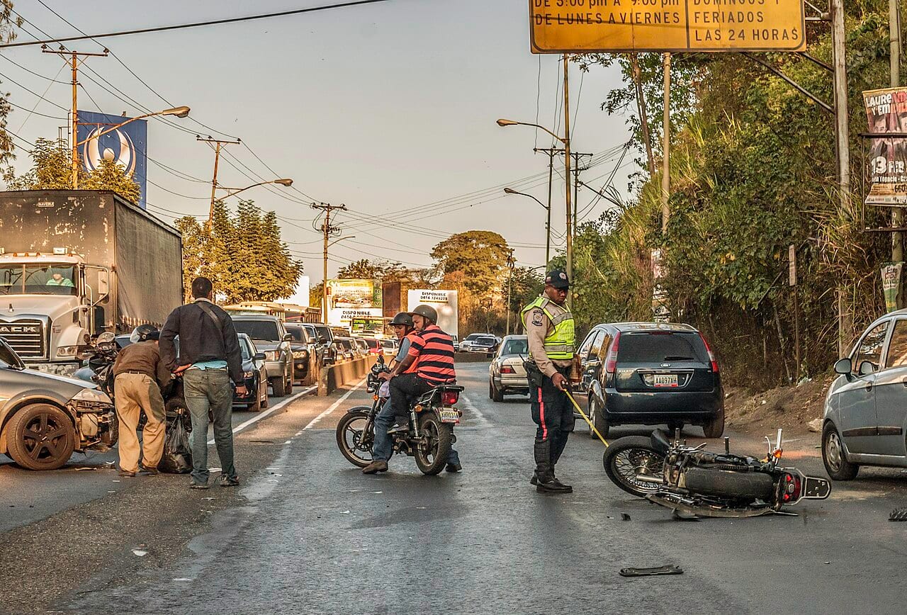 Moto caída en la vía tras un accidente de tránsito, con un agente con chaleco reflectivo y personas atendiendo la escena (foto: Wilfredor, CC0, Wikimedia Commons)